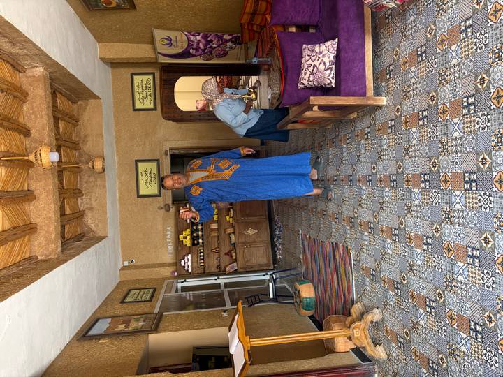 Shop owner in traditional blue robe offers tea inside a decorative carpet and spice store.