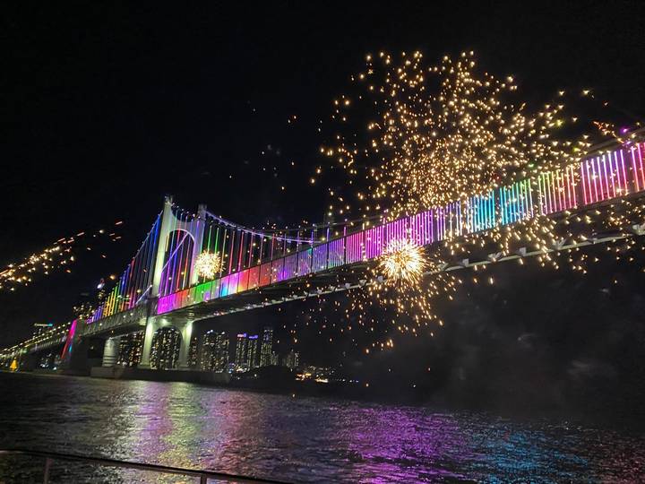 Night fireworks and multicolor light show illuminating a long suspension bridge over the water.