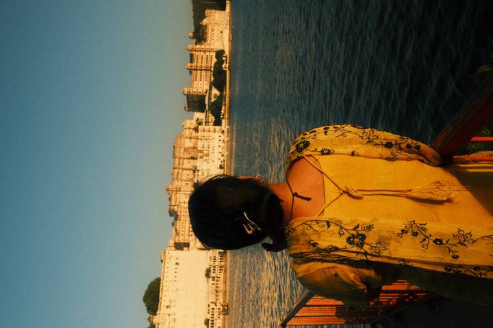 Woman in yellow sari looks across a wide lake toward a sunlit palace complex.