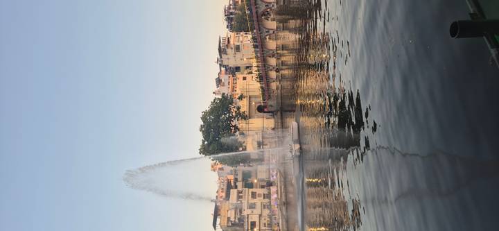Tall water fountain sprays in a city lake beside a busy arched bridge and colourful buildings.