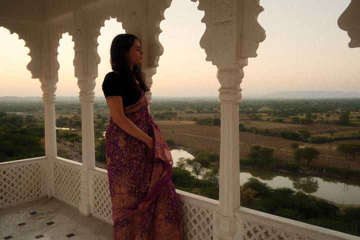 Woman in traditional sari stands on white marble veranda overlooking rural fields at dusk.