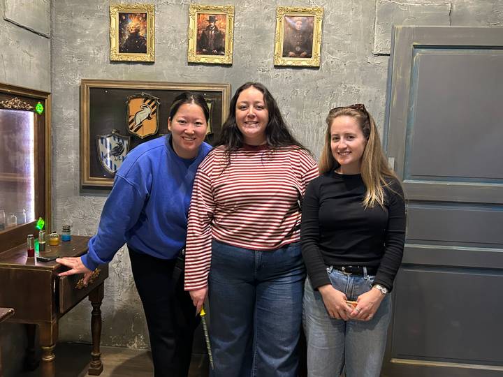 Three women pose together inside a stylish room with framed crests on a textured grey wall.