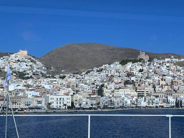 A dense hillside town of white and pastel houses rises from a deep-blue Aegean harbor under clear skies.