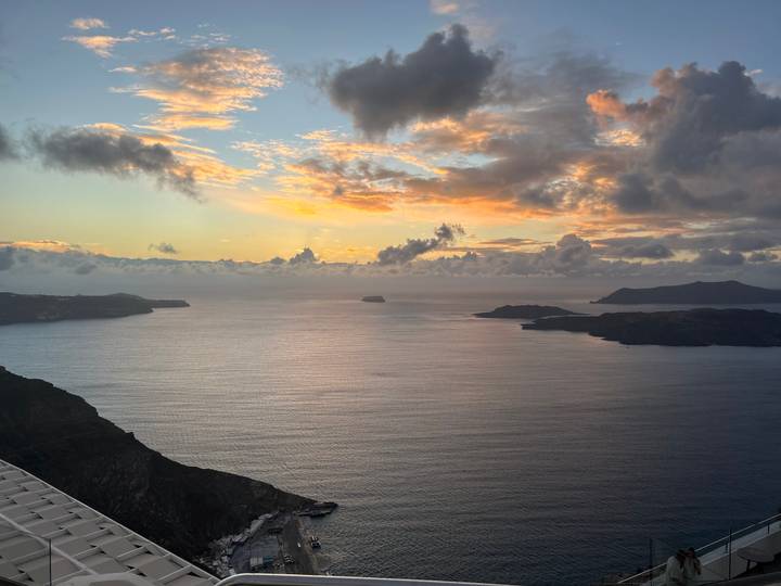 A vibrant sunset colors clouds over the calm Aegean Sea and distant volcanic islets.