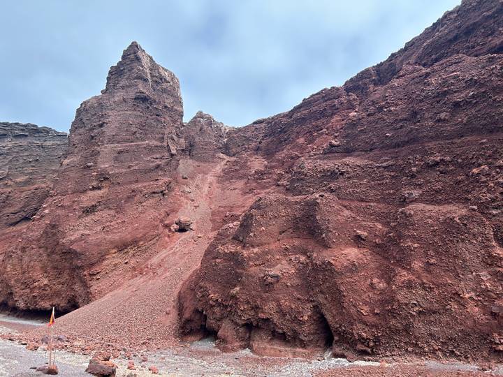 Towering red volcanic cliffs form a rugged canyon beneath a pale blue sky.