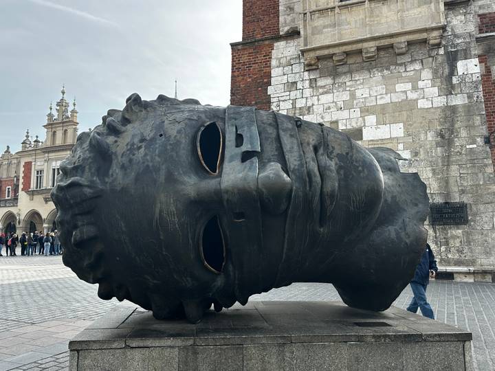 The giant bronze Eros Bendato head sculpture lies sideways in Krakow's main square as visitors walk past.