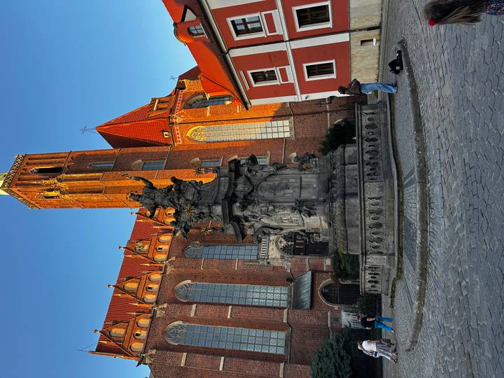 A baroque statue fountain stands before a red-roofed Gothic church in evening light.