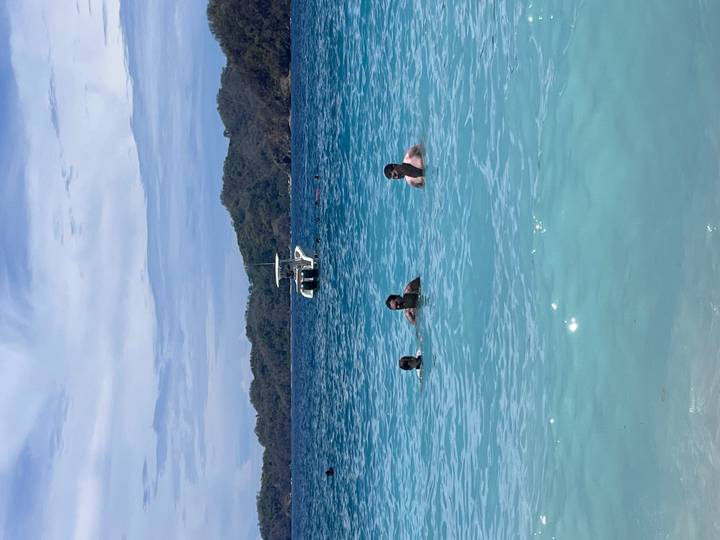 Three swimmers enjoy turquoise tropical waters near a motorboat and forested island backdrop.