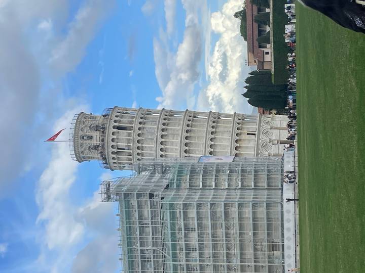 The Leaning Tower of Pisa, partly scaffolded for restoration, tilts distinctively against a blue sky above a green lawn.