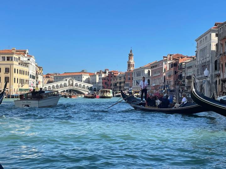 Gondolas glide along Venice’s Grand Canal with the iconic Rialto Bridge and colorful palazzi under a crisp blue sky.