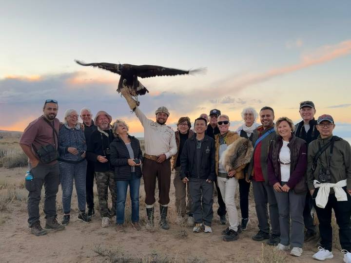 Travellers stand with an eagle hunter who holds a golden eagle at sunset on the steppe.