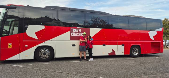 Couple poses beside a large red and white Travel Chaser tour bus in a parking lot.