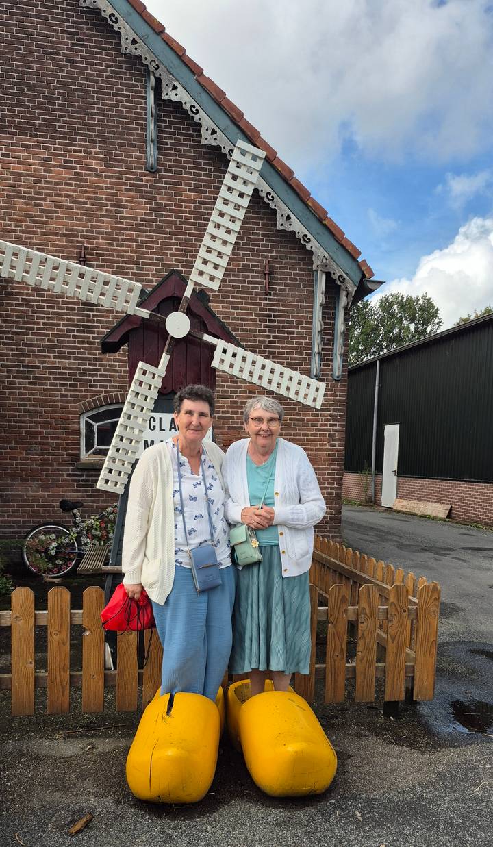 Two older women smile in front of a small white windmill outside a brick building.