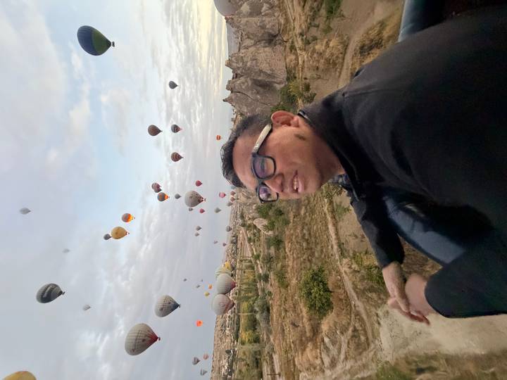 Man leans over balloon basket enjoying sunrise sky filled with dozens of balloons.
