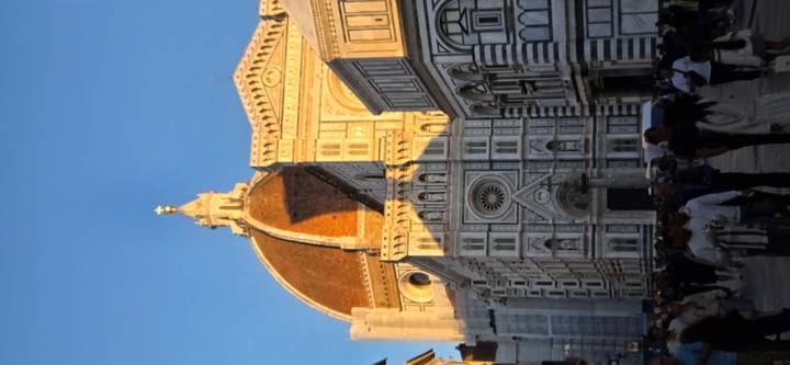 The ornate façade and massive dome of Florence Cathedral glow in warm evening light while crowds stroll the narrow street below.