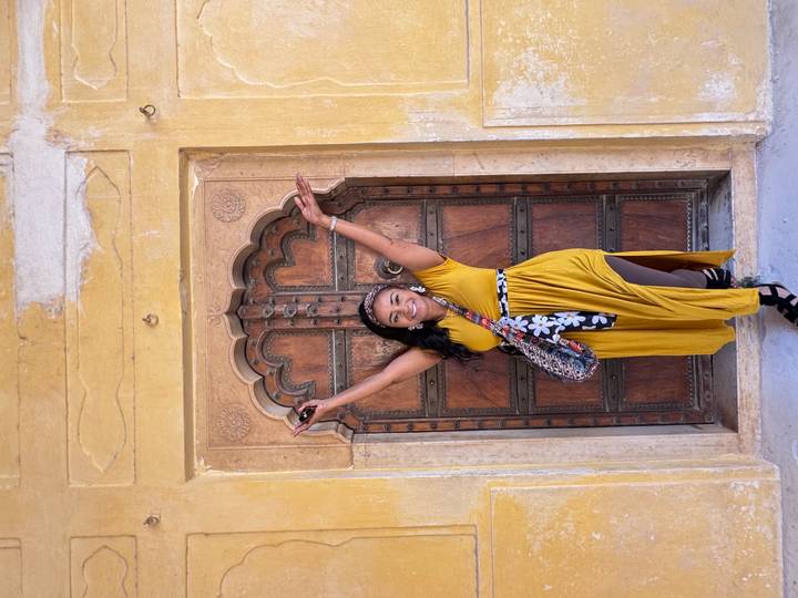 Smiling woman in mustard dress raises arms in front of an ornate arched wooden doorway in a yellow palace wall.