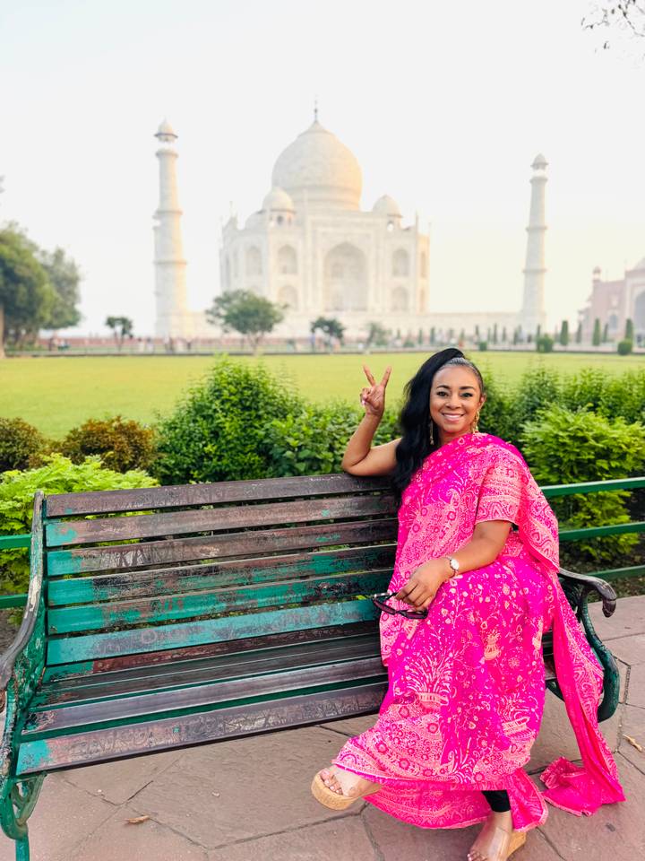 Woman in pink sari sits on a bench flashing a peace sign with the Taj Mahal softly blurred in the background.