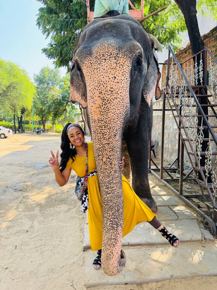 Woman in yellow dress smiles and flashes a peace sign while standing beside an elephant’s trunk.