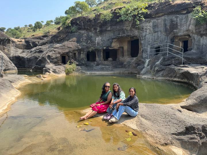 Three women sit on a rocky ledge beside a tranquil natural pool with cave openings in the background.