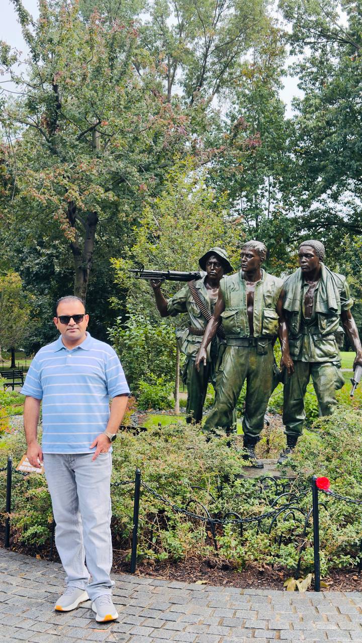 Visitor stands beside the bronze Three Soldiers statue surrounded by greenery in Washington, DC