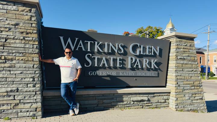 Traveler poses beside the large stone sign for Watkins Glen State Park on a sunny day