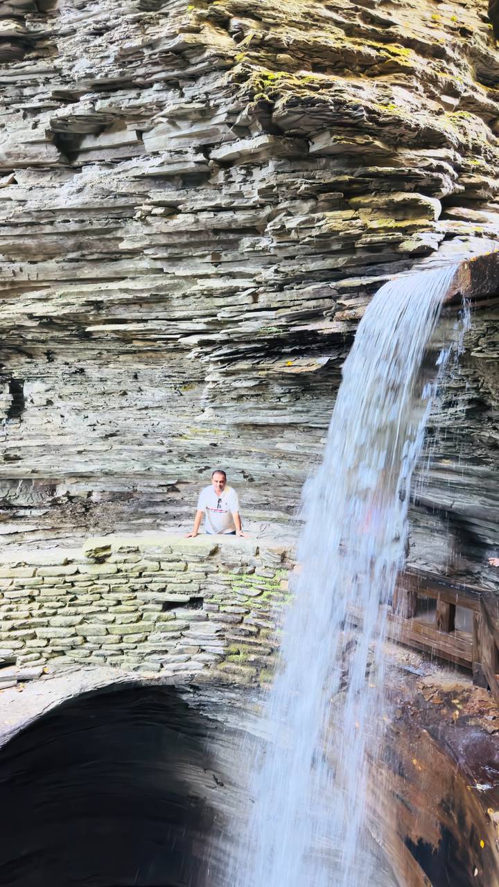 Man kneels behind a curtain of waterfall inside a rocky gorge with layered stone walls