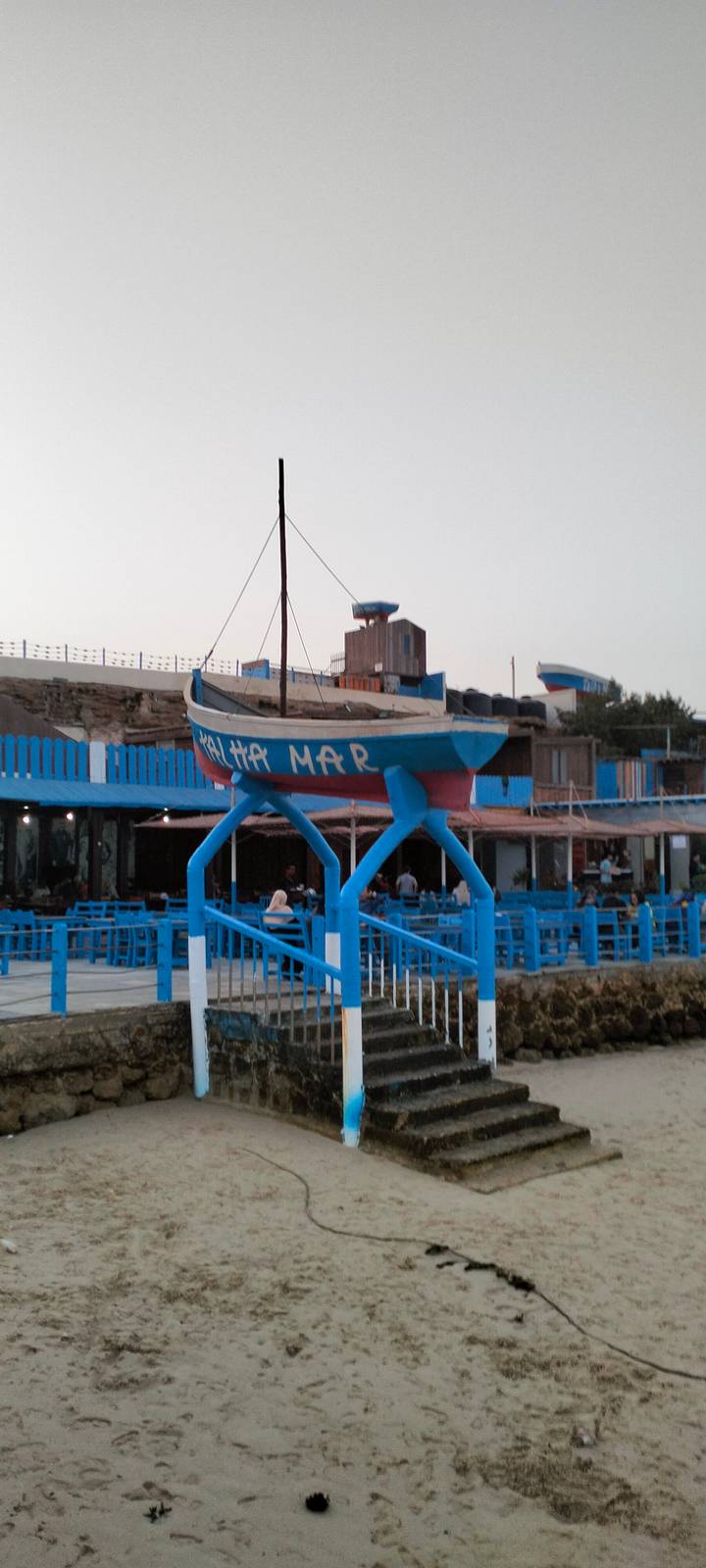Blue and red boat sculpture elevated on stilts in front of a seaside café complex, image slightly blurry