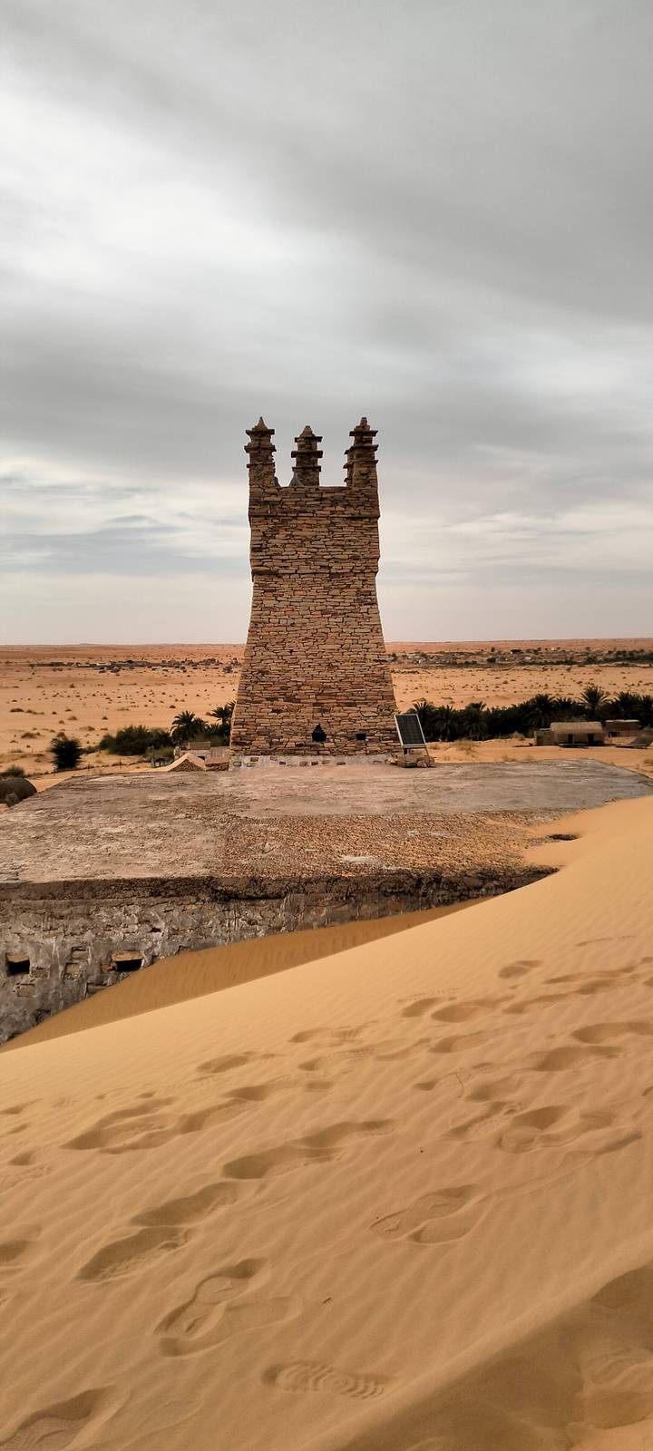 Stone tower rising above desert dunes and distant oasis vegetation under a hazy sky