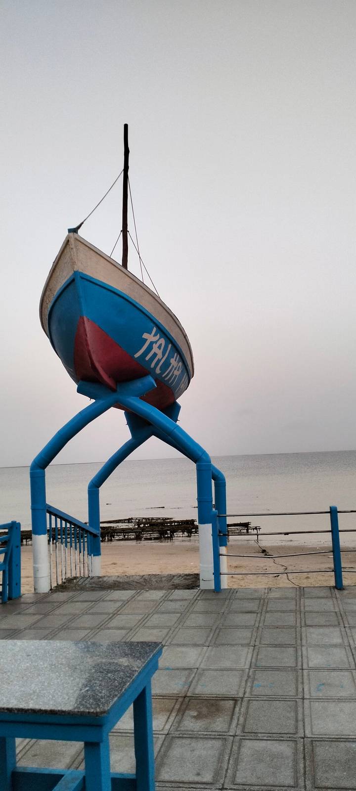Blue boat sculpture on stilts set against an empty horizon of calm sea and pale sky