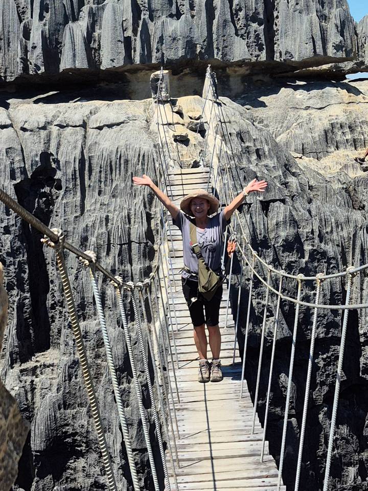 Traveller with arms raised crosses a narrow suspended wooden bridge between jagged grey limestone pinnacles.