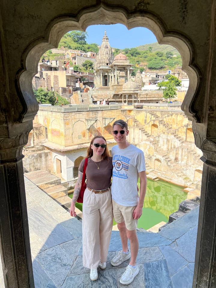 Smiling couple standing inside ornate yellow stepwell with carved arches and green water in Jaipur.