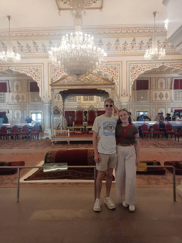 Visitors pose in opulent hall of Jaipur City Palace featuring ornate silver throne, mirrors and archways.