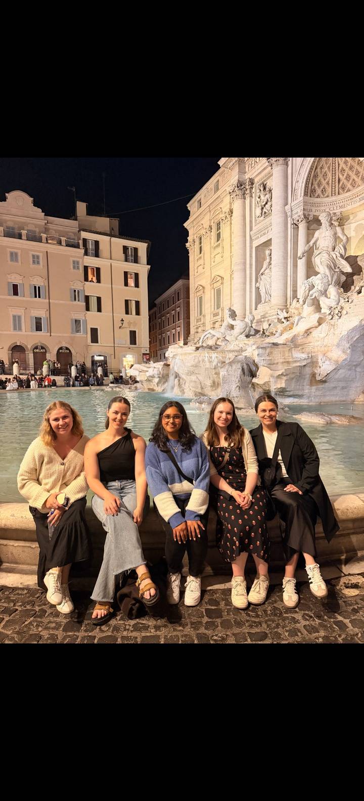 Five friends pose on the edge of the Trevi Fountain with softly lit statues behind them.