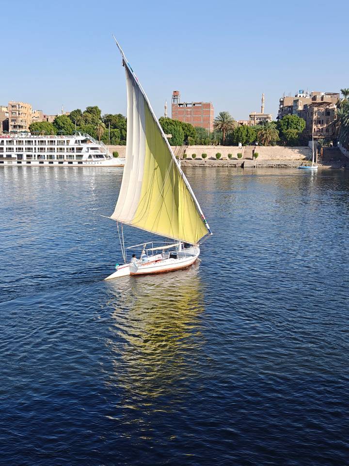 A traditional felucca with a large cream-colored sail glides over the calm blue Nile River.