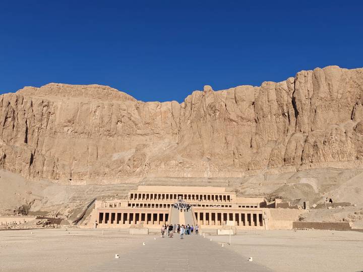 Temple of Hatshepsut stands at the base of towering desert cliffs with visitors walking up its ramps.