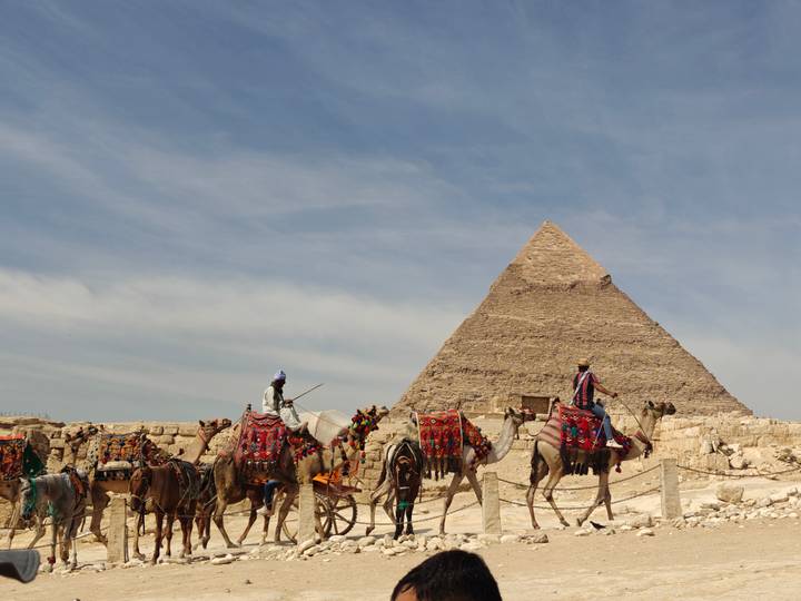 Camel riders in colorful blankets pass in front of the Great Pyramid under a lightly clouded sky.