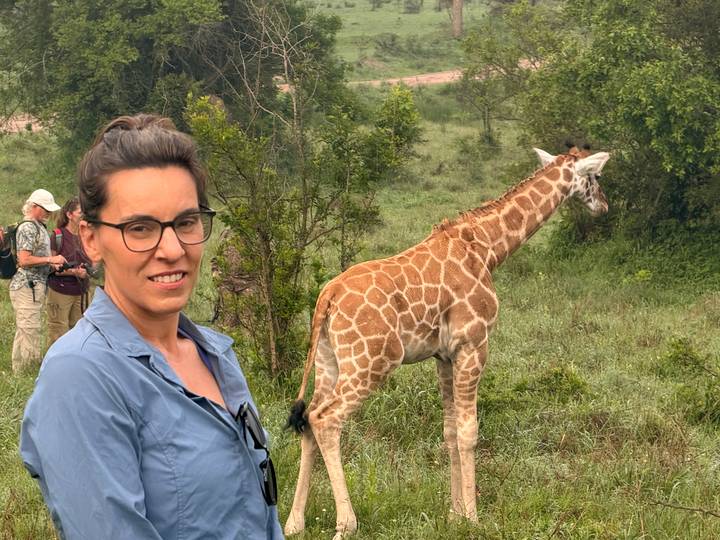 A smiling woman poses near a young giraffe while fellow travelers observe wildlife behind.