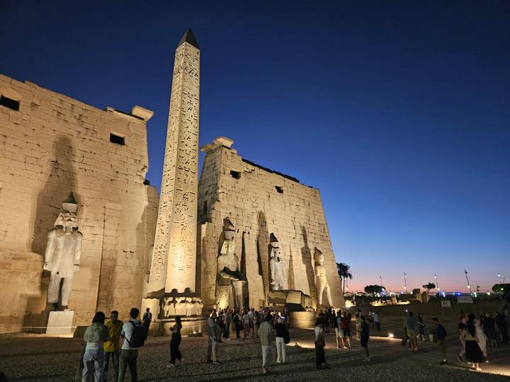 Illuminated statues, obelisk and colonnade of Luxor Temple against a blue twilight sky.