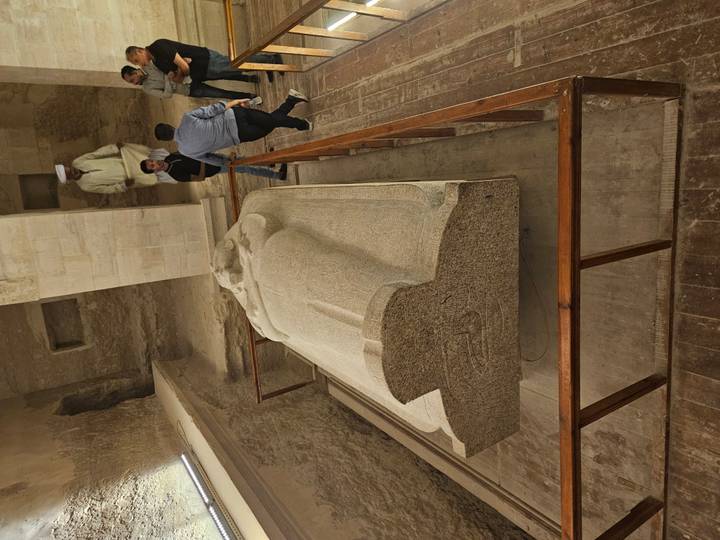 Granite sarcophagus displayed in an underground Egyptian tomb chamber while three visitors observe behind a wooden barrier.