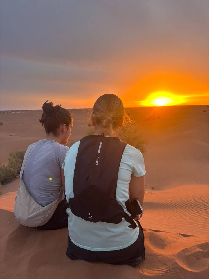 Two travelers sit on a dune watching fiery sunset over rolling Sahara sands.