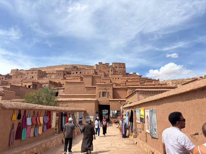 Adobe-brick ksar of Aït Benhaddou rises above market stalls and tourists under blue sky.