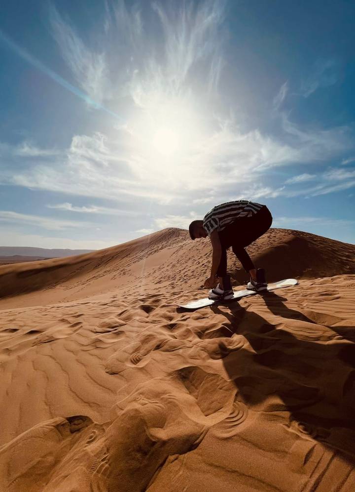 Snowboarder crouches on golden dune preparing to sandboard beneath bright desert sun.