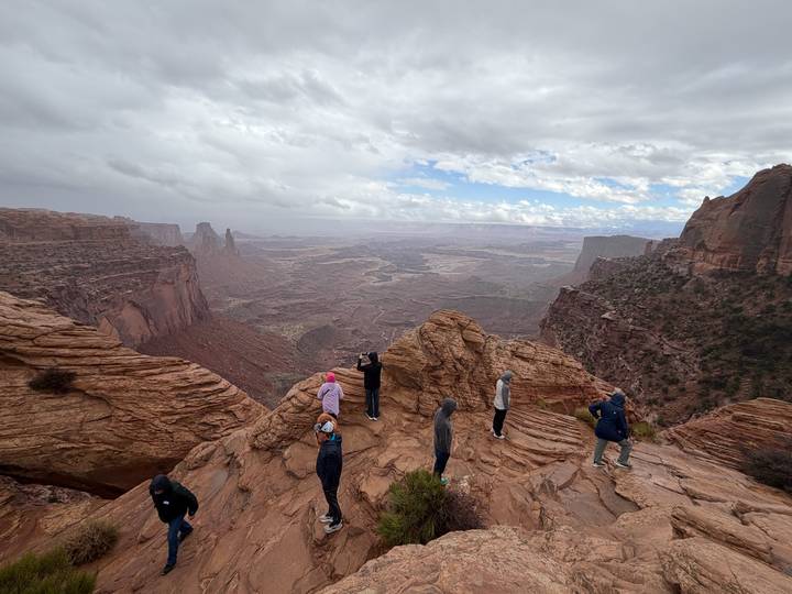 Hikers stand on wind-carved sandstone overlooking Canyonlands’ vast canyon landscape under a cloudy sky.