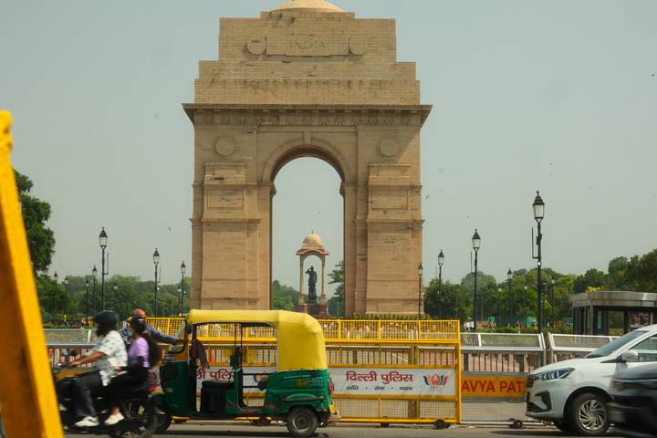India Gate monument framed by busy city traffic and hazy sky, with auto-rickshaw in foreground.
