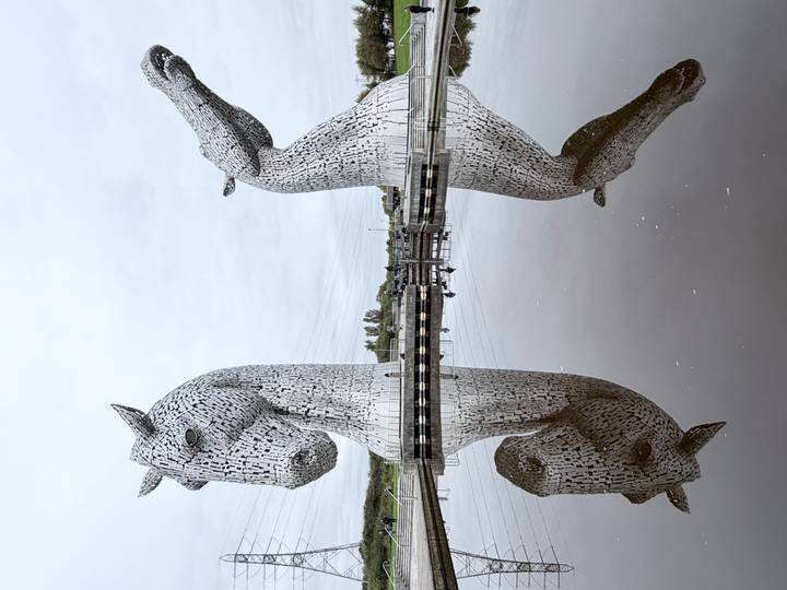 Massive steel horse head sculptures known as The Kelpies reflected perfectly in still water under a grey sky.