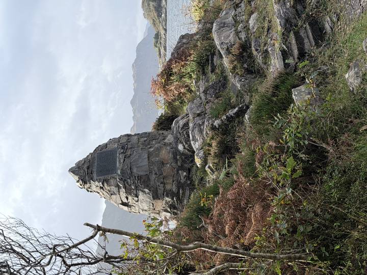 Stone cairn memorial on rocky terrain surrounded by ferns and shrubs with distant mountains.