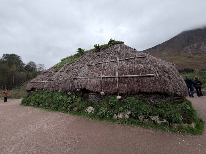 Traditional thatched blackhouse structure with visitors standing nearby under overcast skies and misty mountains.