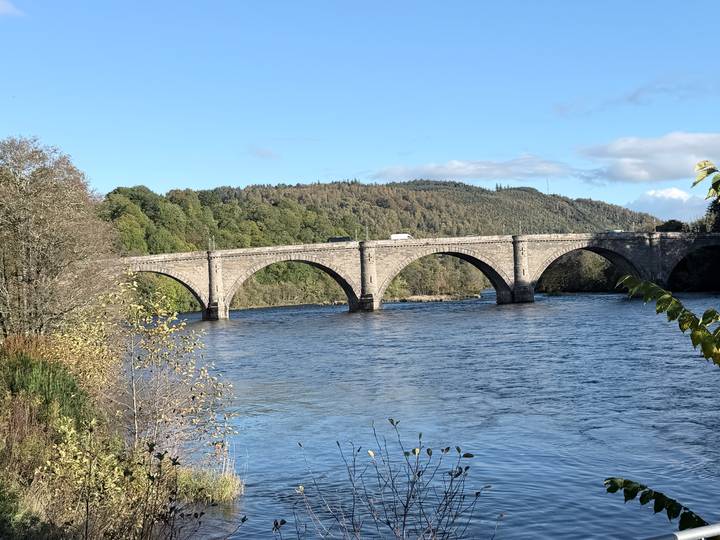 Historic stone arch bridge spanning a wide river bordered by forested hills under clear blue skies.