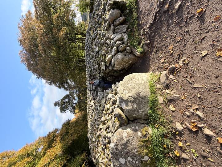 Visitor standing inside an ancient circular stone cairn surrounded by trees under blue skies.