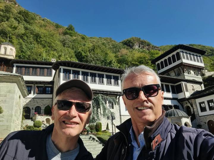 Two travellers taking a selfie in front of an ornate Macedonian monastery complex set against forested hills.